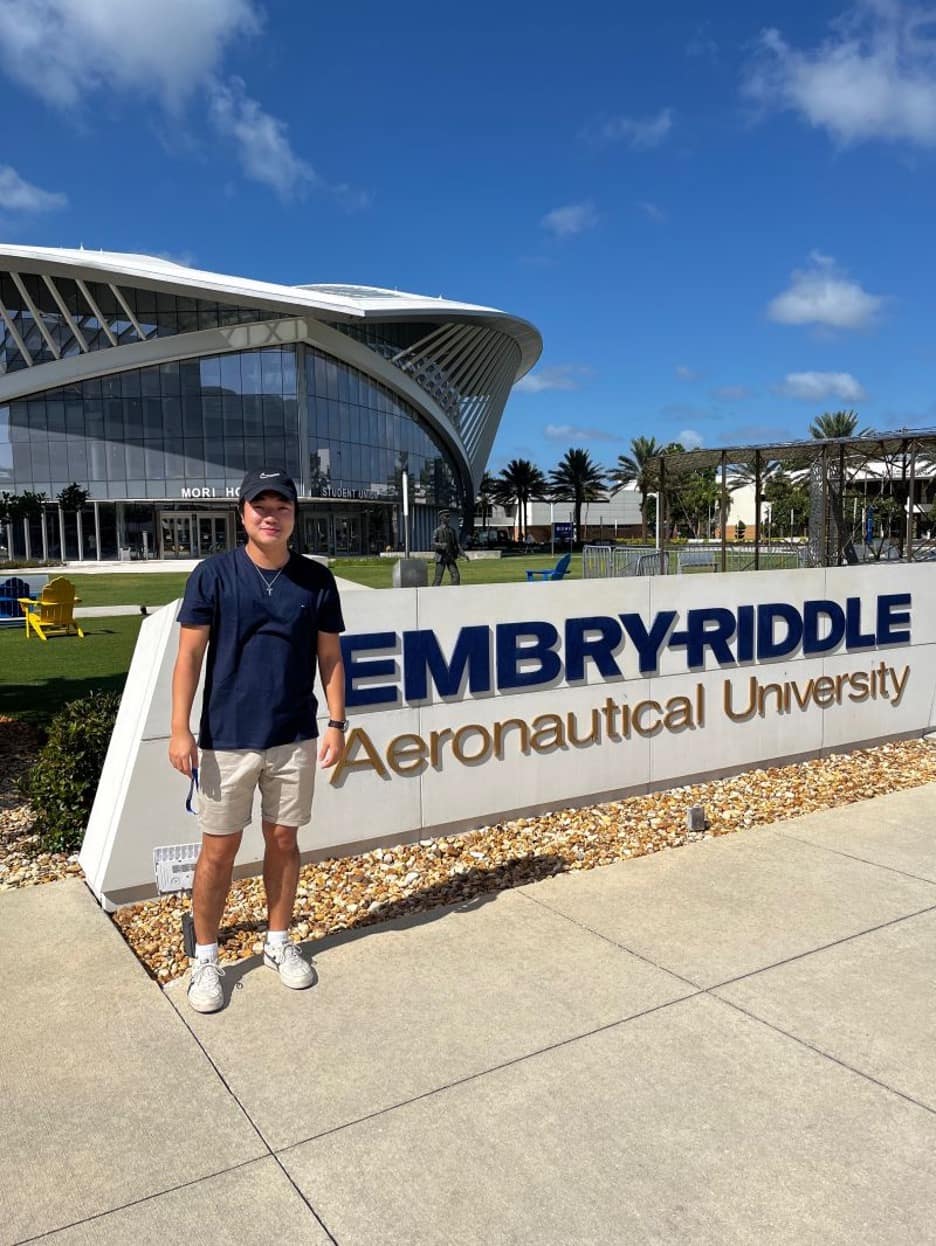 Mark Jacob standing in front of the Embry-Riddle monument sign with the sweeping roofline of the student union and landscaped grounds in the background.