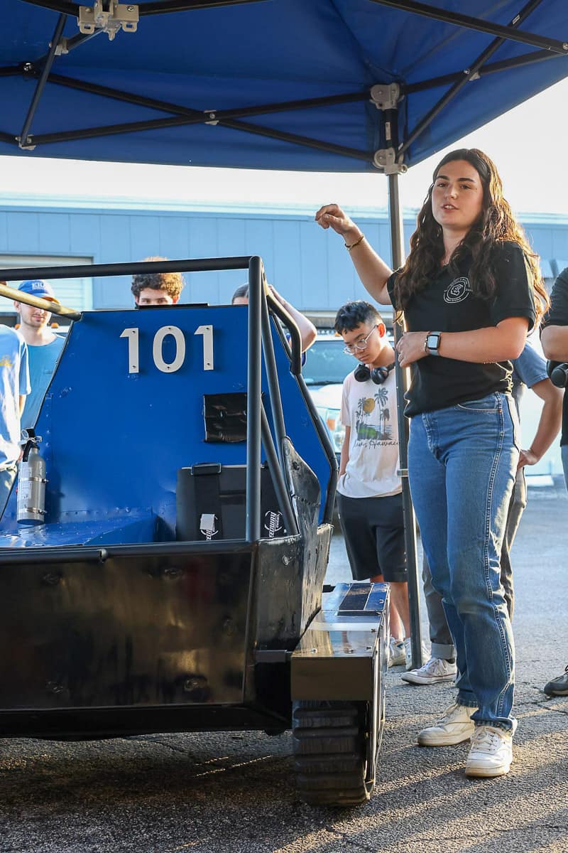 Woman stands near tracked Baja vehicle.