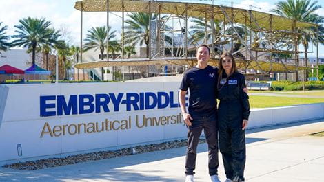 Embry-Riddle alumna Maria Noel de Castro Campos and her fiance, David, pose on campus in front of a wall with the University's name.