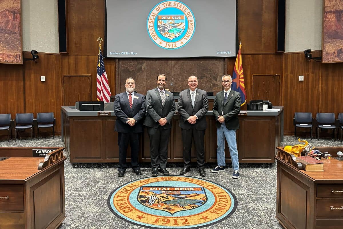Four men stand in a formal room with the Arizona state seal on the floor and wall. They're dressed in suits, except one in jeans. The tone is official.