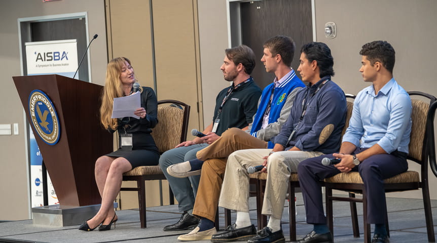 Four students sit in a row on dais while they are asked questions by a professor holding a microphone