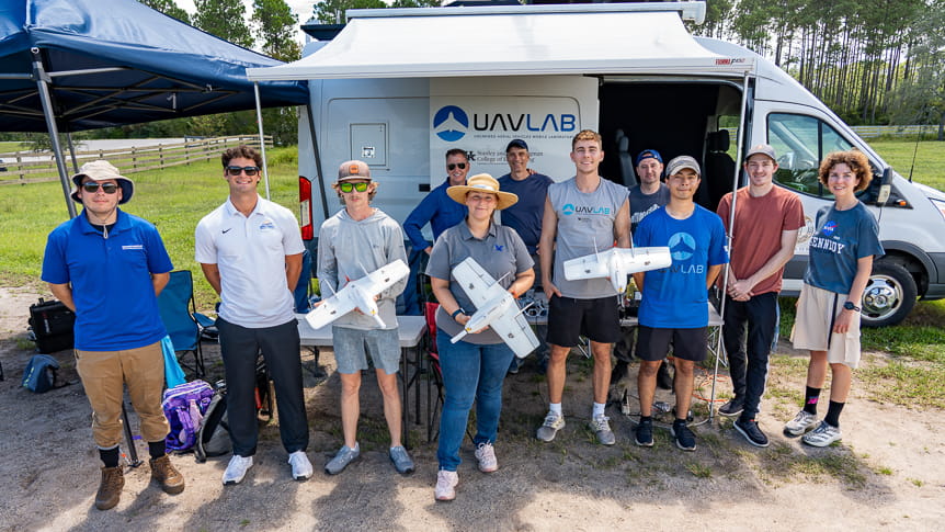 Group of twelve people standing in front of UAV lab mobile van holding drone 