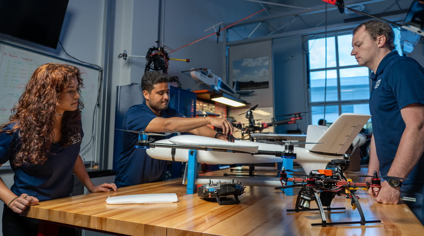 Three students and a table on which sits a small, white drone