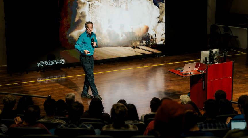 Former astronaut Jim Wetherbee on a stage in front of an image of the space shuttle taking off. 