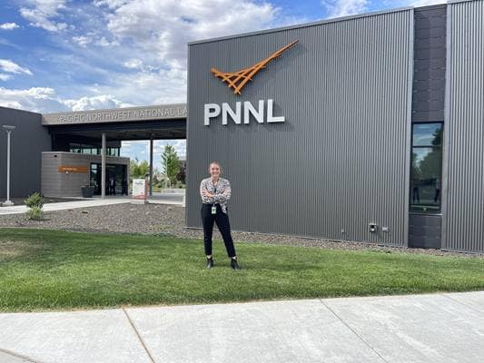 A person standing on grass in front of a gray building which has the letters PNNL on the outside. 