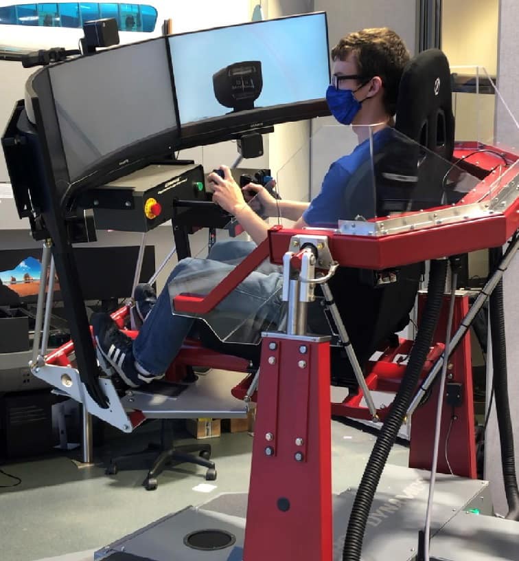 Student operating a multi-axis flight simulator with triple curved monitors, joystick, throttle controls, and rudder pedals, seated in a motion platform cockpit rig with exposed actuators and safety shield in a lab.