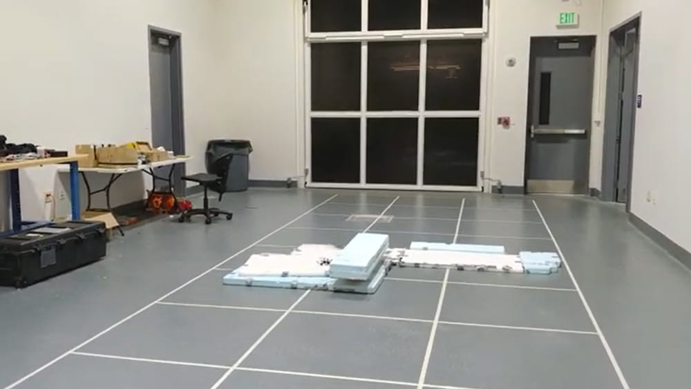 Drones laid out on a marked floor in a high-bay lab, with tool carts, storage bins, and a large roll-up door in the background.