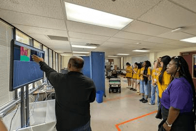 A group of students stand in a classroom as a instructor presents and shows a graphic on a large monitor