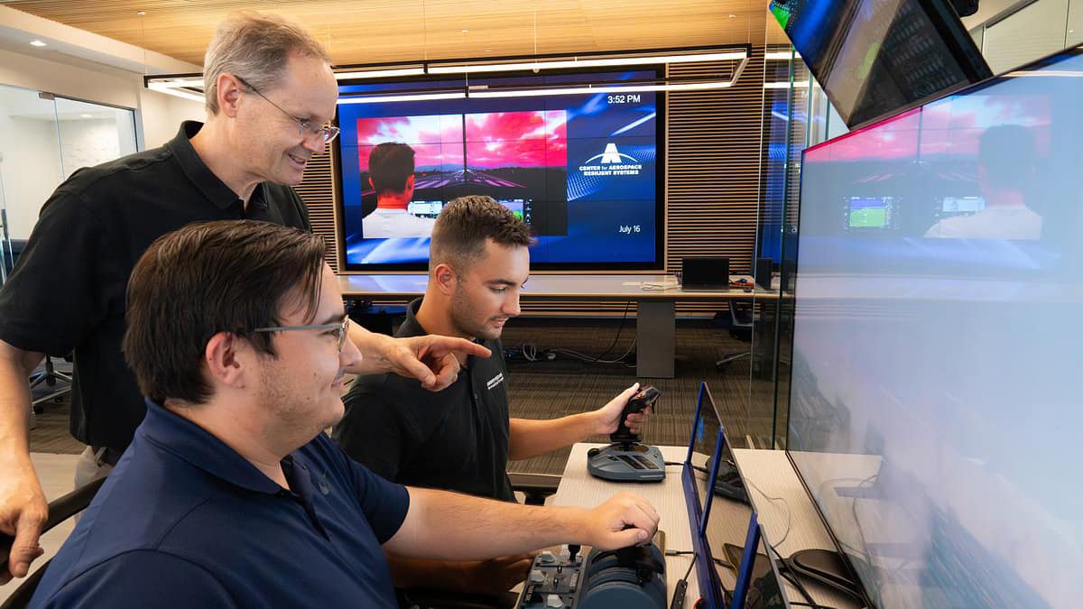 A professor watches two students who are using a flight simulator.