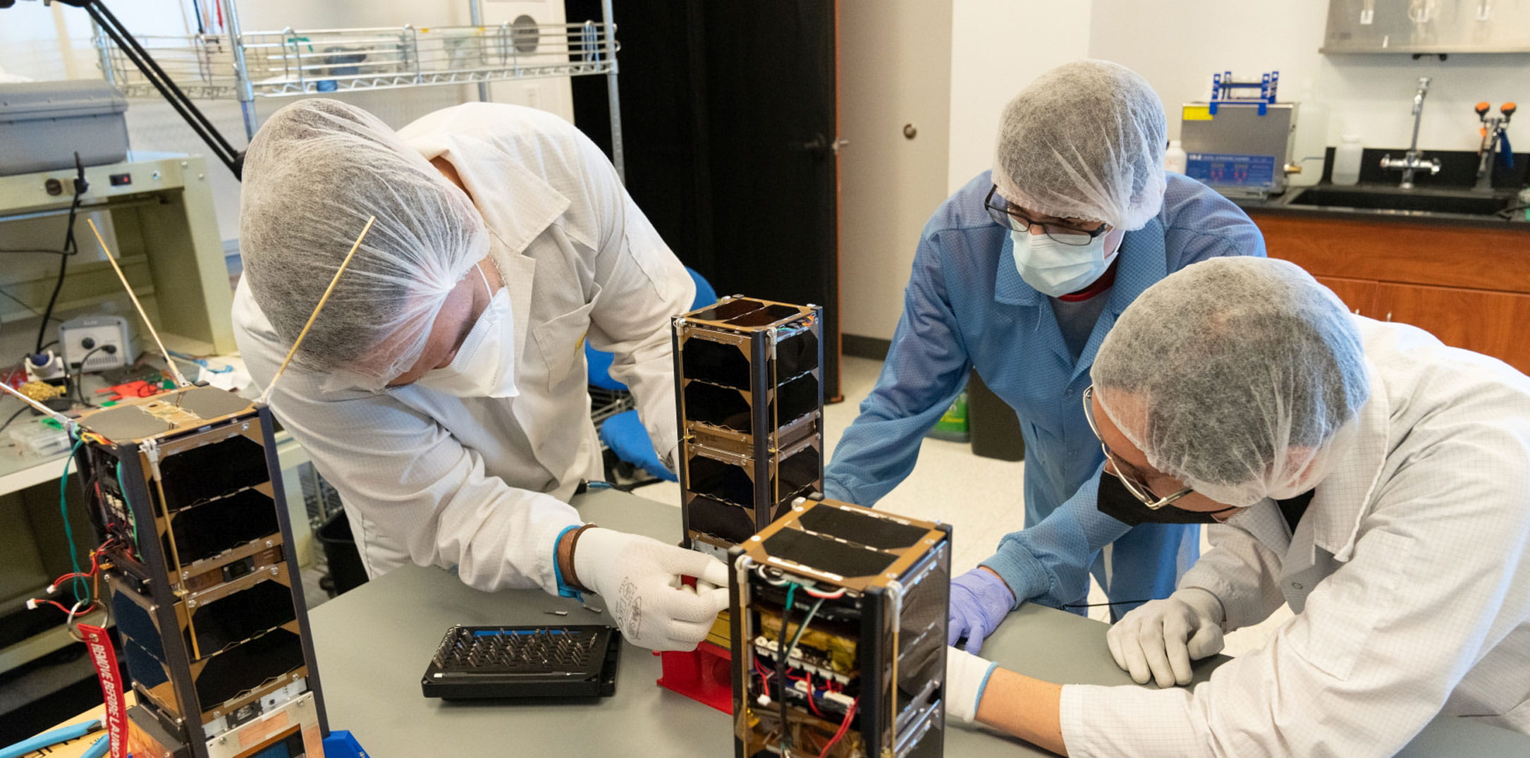 Three students with masks and lab coats work on a project.