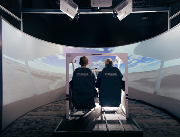 “Two people sit side by side in simulator seats, facing a large curved panoramic screen inside a dark training room.