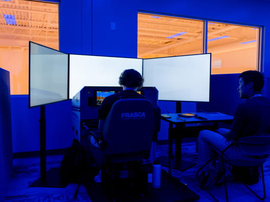 Two people sit at a workstation in a dim, blue-lit room, facing multiple computer monitors.