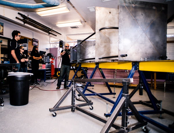 A workshop with a large metal chassis frame mounted on adjustable jigs, welding equipment nearby, and several people working in the background.