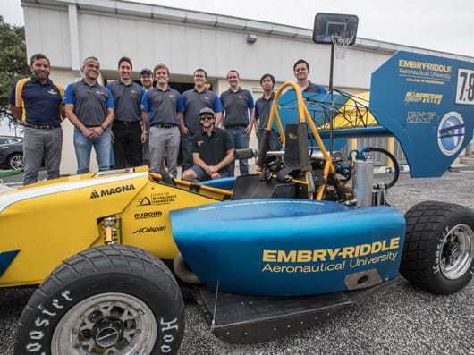 A group of people standing behind an Embry-Riddle race car outdoors.