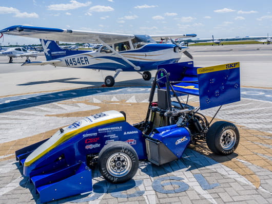 A blue race car parked outdoors in front of a small Embry-Riddle airplane.