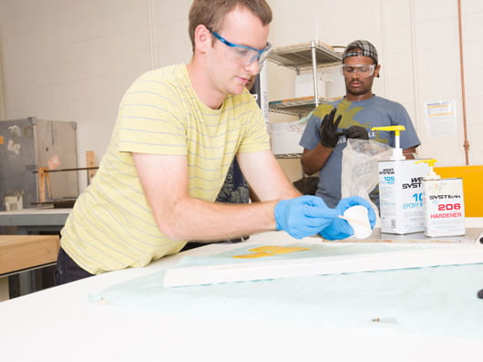Two people wearing safety glasses and gloves work in a workshop.