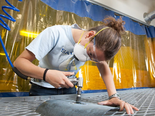 A person wearing safety glasses and a mask, working in a workshop.