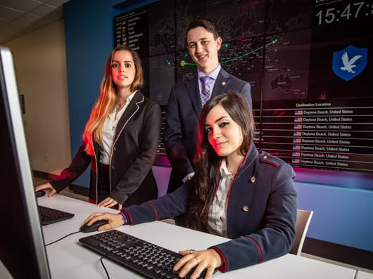 Three students in business attire collaborate at a computer workstation, with one seated and two standing nearby