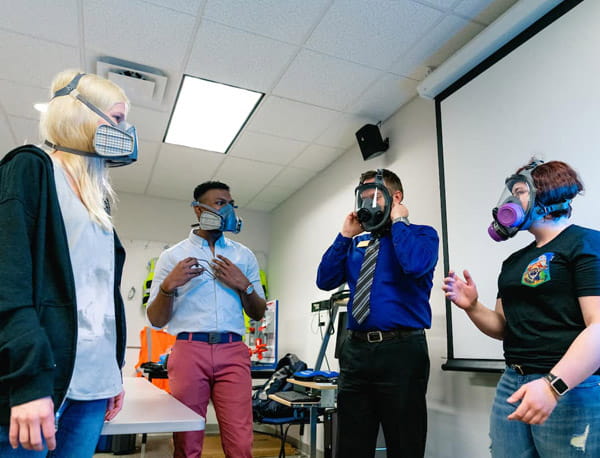 Four people wearing gas masks in a classroom setting.