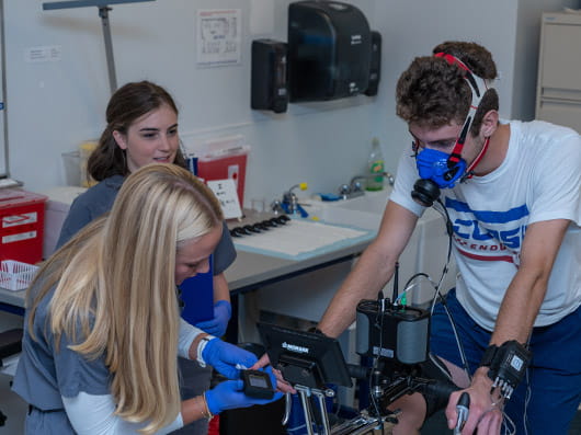 A person pedals a stationary bike while wearing a mask, as two clinicians monitor their health.