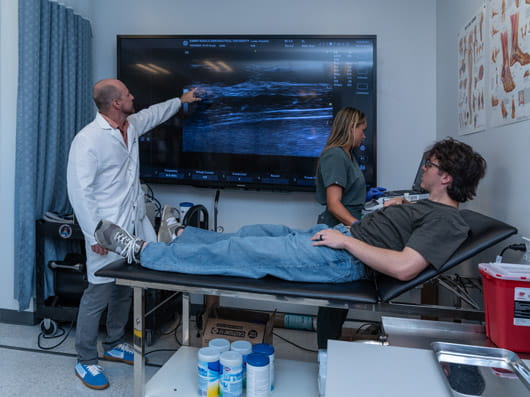 A patient lies on an exam table while two clinicians review an ultrasound image displayed on a large monitor, with one clinician pointing at the screen.