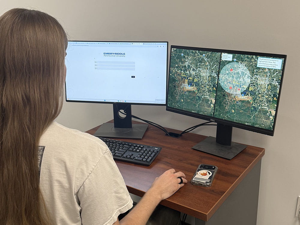 A student sits at a small desk working on a computer with two monitors. One monitor has a map software opened on it.