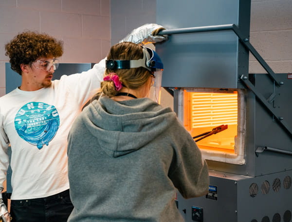 Two people wearing safety equipment while operating a kiln.