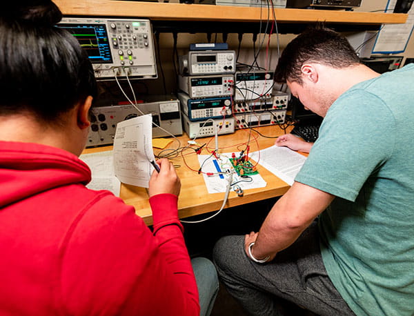 Students read notes at a desk, surrounded by devices and wires.