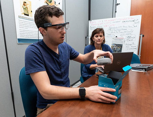 A student wearing eye tracking hardware unboxes a product while a professor observes.
