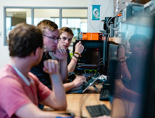 Students converse at a desk in front of many computer screens.