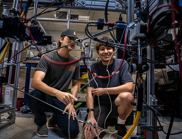 Two students crouch and look at wires and cables hooked up to equipment.