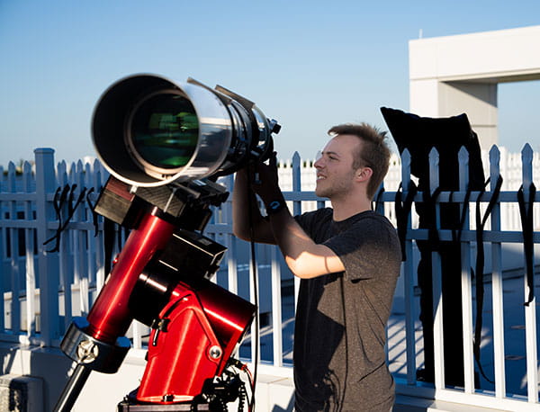 A male tudent adjusts a large telescope outside.