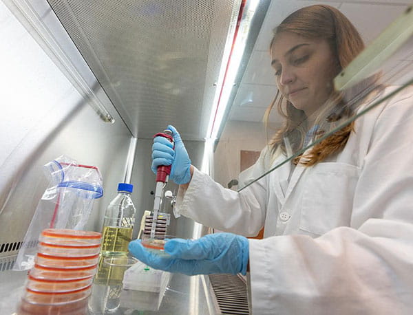 A student in lab clothes puts material in a petri dish.