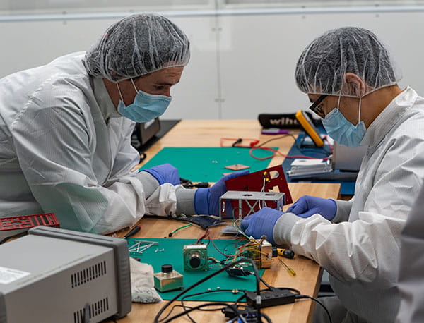 Two people in protective equipment work at a desk putting together a small aparatus.