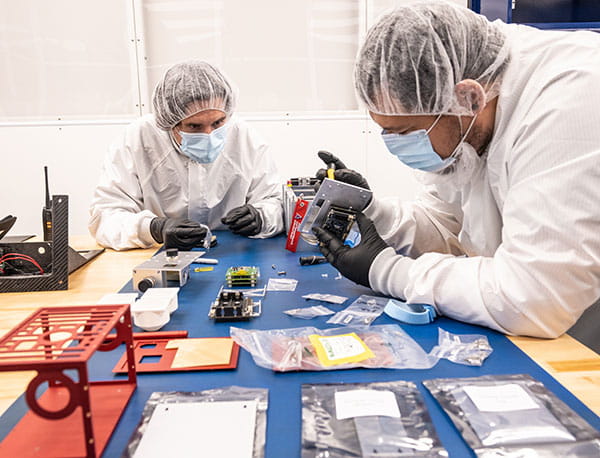 Two students in protective equipment put together various pieces of an aparatus on a desk.