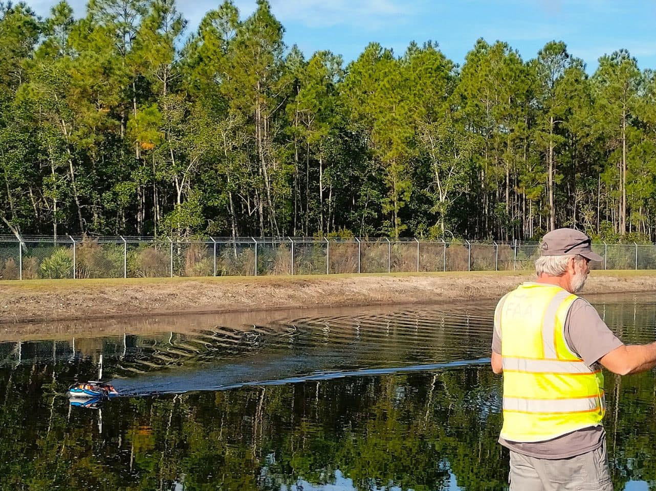 Individuals stand on the bank of a pond where an uncrewed vehicle glides across the water