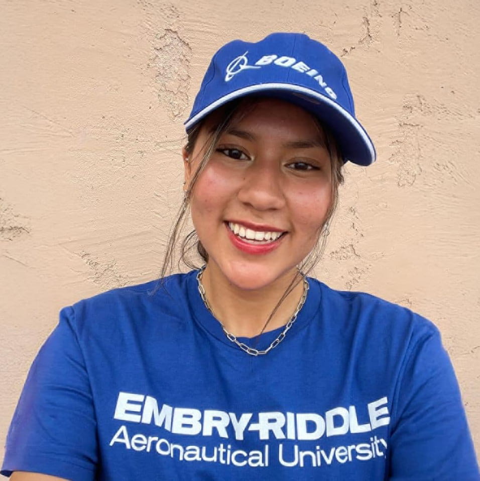 Embry-Riddle student, Camille Hawkins, stands against a wall in a blue Embry-Riddle shirt and Boeing hat