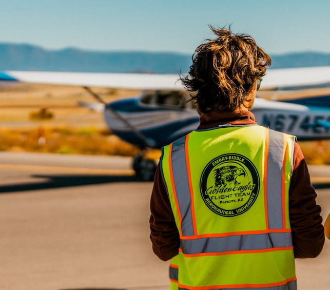 An Embry-Riddle student in a yellow safety vest stands on the tarmac observing a small plane going by