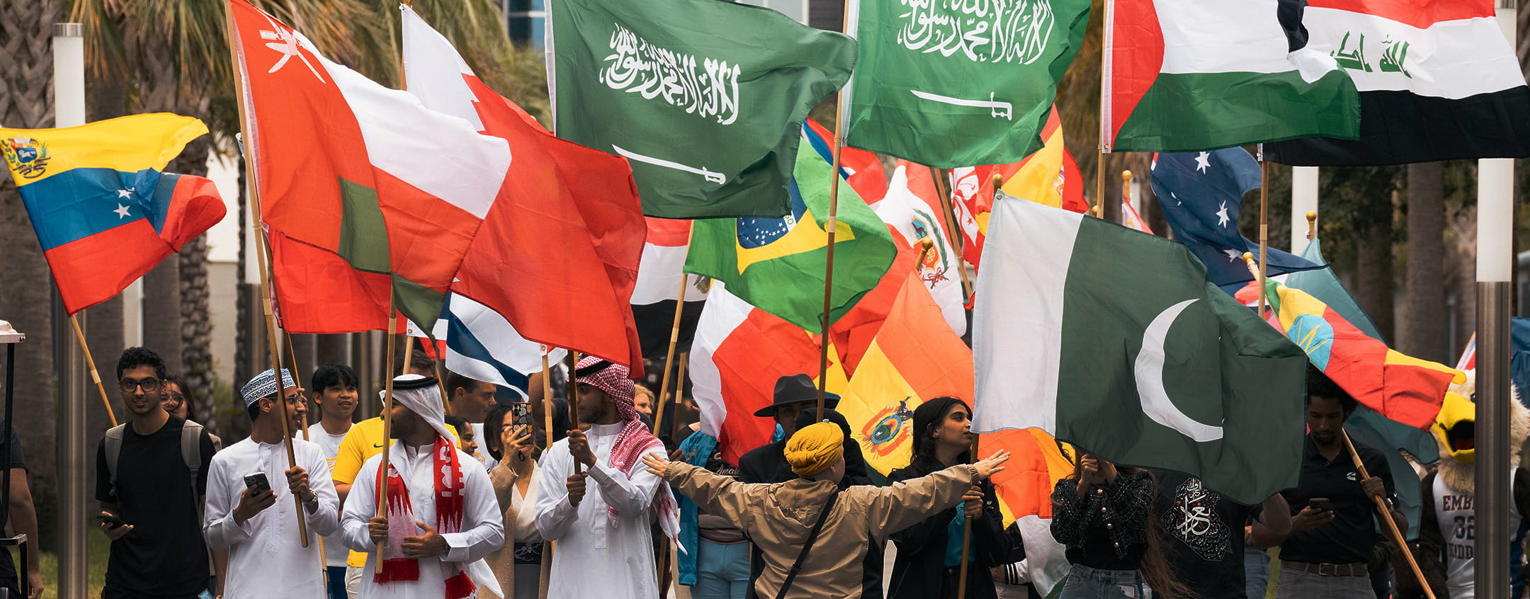 Students carry large national flags including Saudi Arabia, Pakistan, Brazil, Oman and Bahrain during an outdoor international parade on campus, with palm trees and a crowd of participants walking together behind them.
