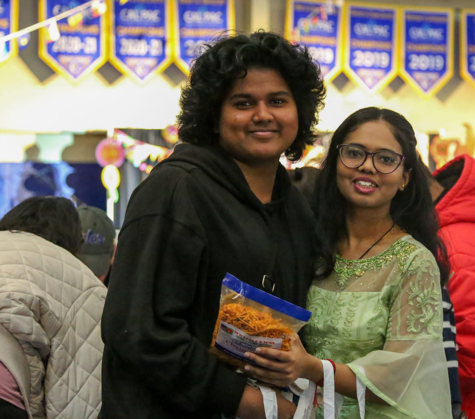 Two students pose together at an indoor international festival, with strings of small international flags overhead and Cal Pac championship banners lining the gymnasium wall in the background.