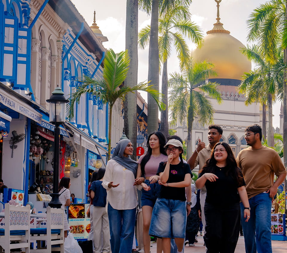 Students walk through a street market lined with shophouses toward a large gold-domed mosque, Sultan Mosque in Singapore’s Kampong Glam district, with palm trees and storefront signs visible along the walkway.