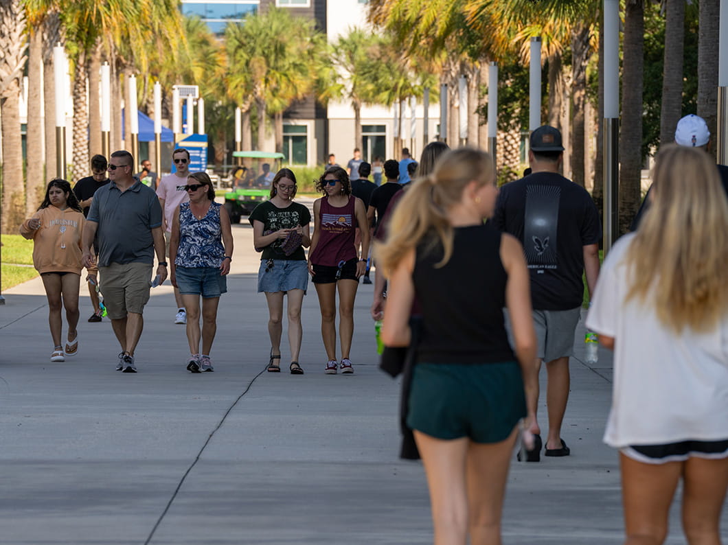 Students walking along palm-lined campus pedestrian walkway with academic buildings and golf cart in background.