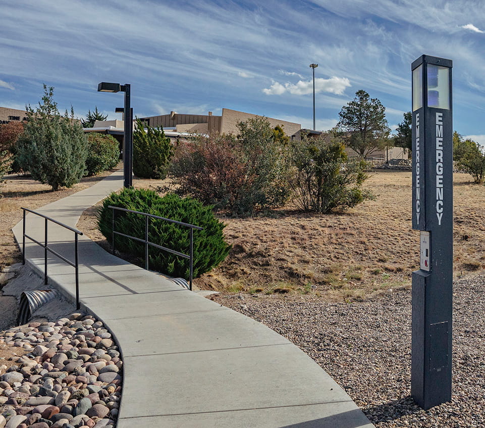 Emergency call box labeled 'Emergency' beside accessible campus walkway with handrails, desert landscaping, and academic building in background.