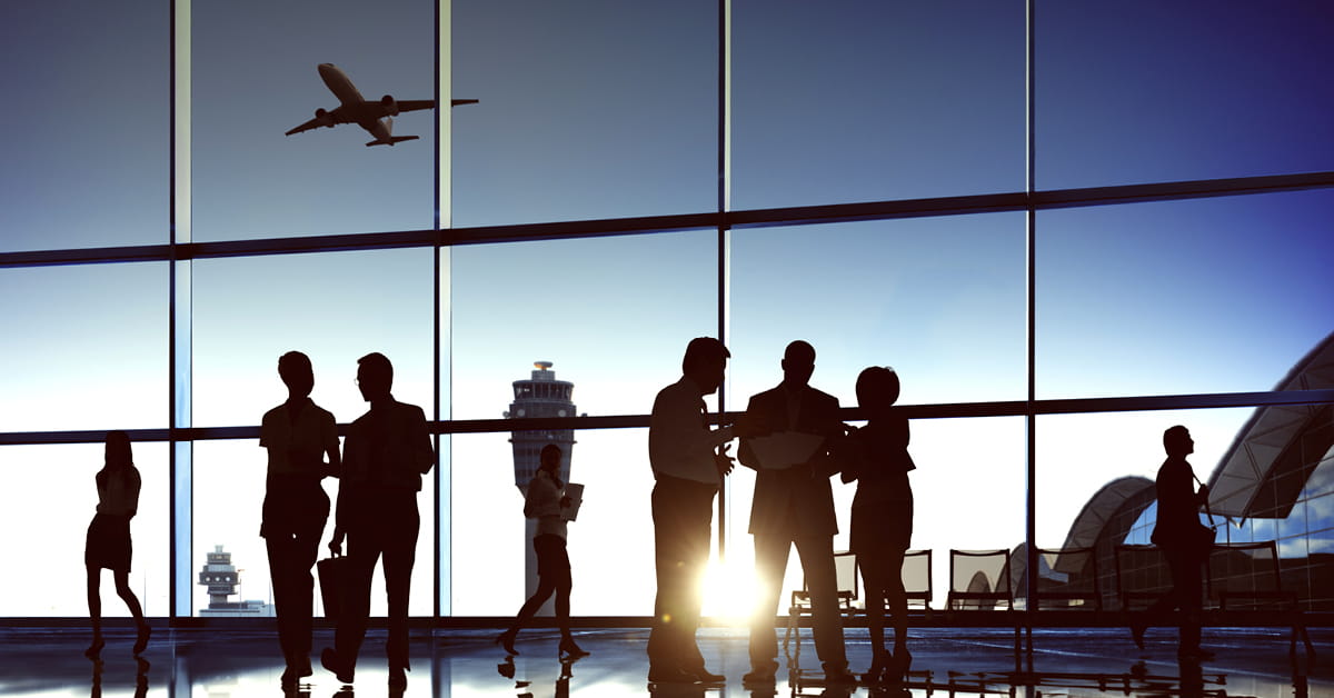 Several people in silhouette standing in front of a bank of windows at sunset, a jet taking off in the middle distance.