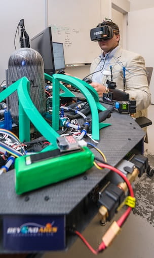 Student wearing a virtual reality headset operates a carbon fiber–framed robotic system with compressed gas cylinder, solenoid valves, wiring harnesses, and green protective brackets in a lab setting.