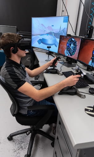 Student wearing a virtual reality headset operates dual flight control sticks at a workstation with three monitors displaying aircraft simulation graphics and a spacecraft model, in a computer lab environment.