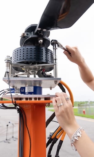Close view of a multirotor drone propulsion test stand with mounted brushless motor, propeller hub, perforated shroud, wiring, and torque arm on an orange support column, as a technician tightens a bolt with a wrench outdoors.
