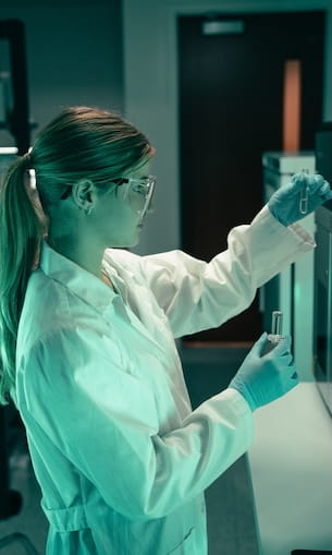 Student wearing safety glasses and a lab coat transfers liquid between test tubes while working at a laboratory bench with analytical equipment.