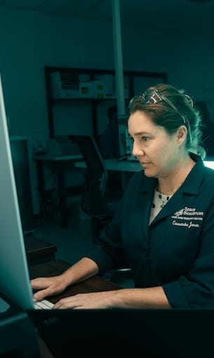 Faculty member wearing a lab coat labeled Space and Atmospheric Research works at a desktop computer in a laboratory office setting.