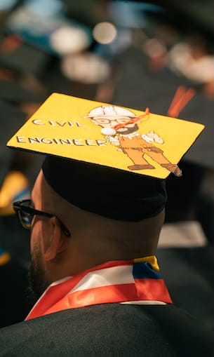 Graduation cap decorated with a cartoon construction worker and the words Civil Engineer, worn by a graduate during commencement ceremony.